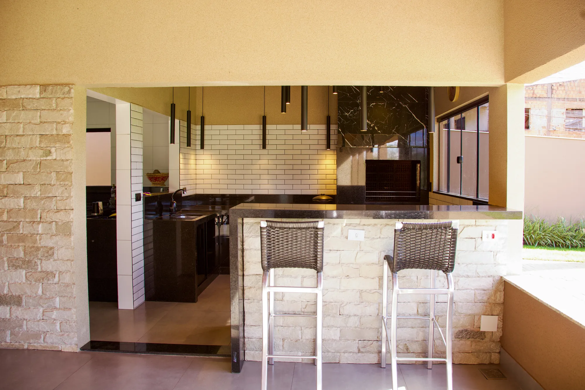 Área gourmet com balcão de granito preto, banquetas, pendentes e churrasqueira ao fundo